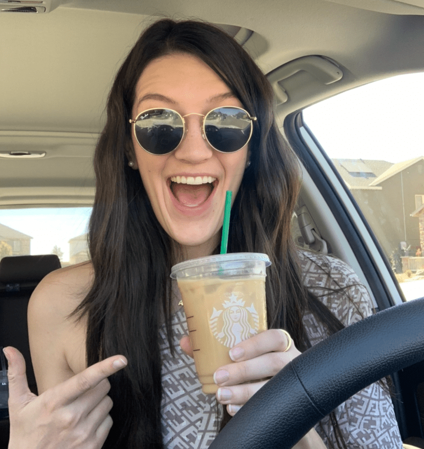 Young white woman with long dark hair and sunglasses, sitting in a car, very excited about an iced coffee